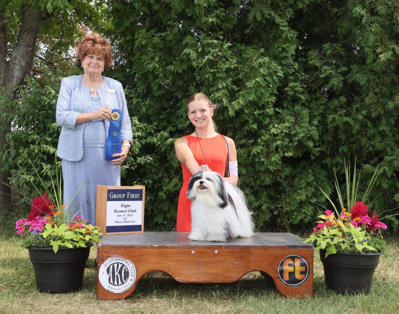 Two women standing next to a white dog with a ribbon.