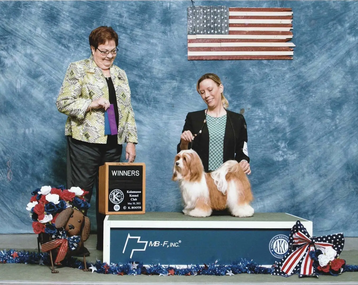 Two women standing next to a Havanese dog in a show ring.