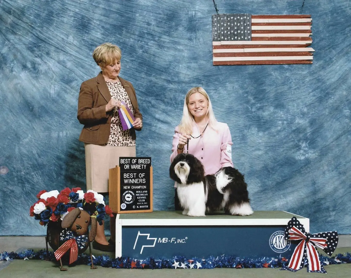 A woman and a Havanese puppy standing next to an American flag.