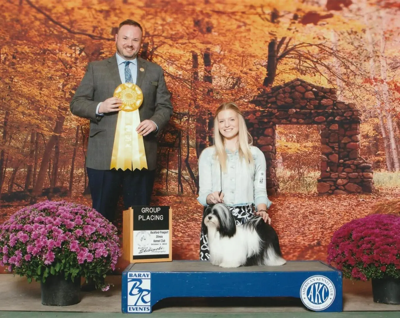 A man and woman standing next to a Havanese dog on a stage, showcasing their champion grand champion Havanese puppies.