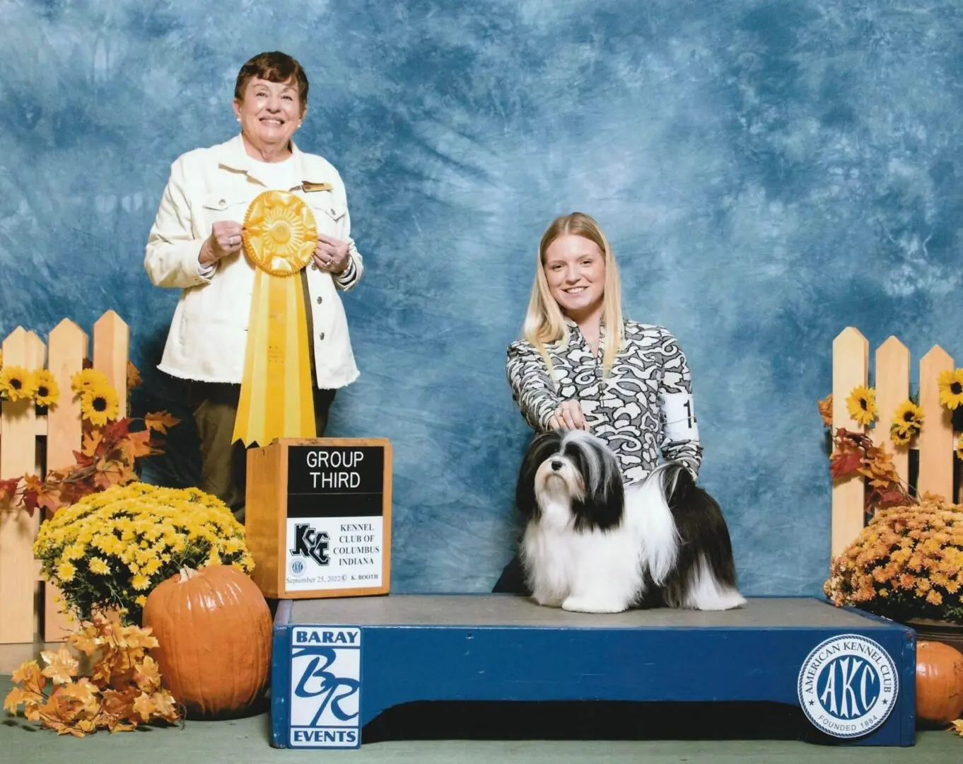 Two women posing with a shih tzu puppy in front of a pumpkin.