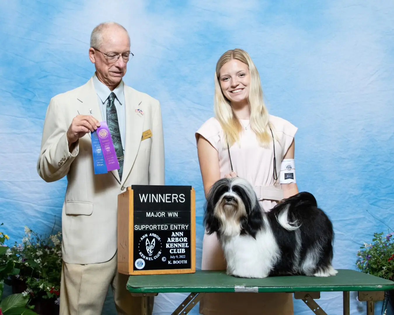 A man and woman standing next to a Havanese dog with a ribbon.