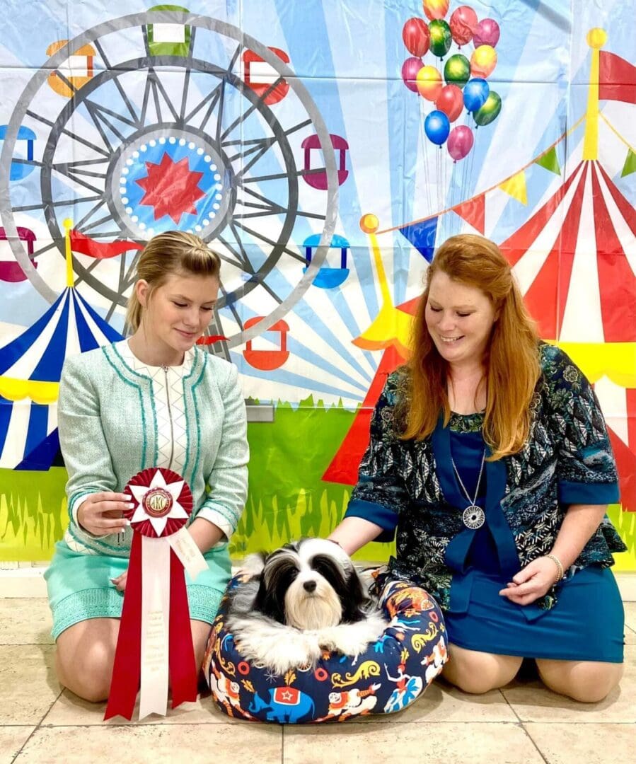 Two women posing with a Havanese dog in front of a ferris wheel.