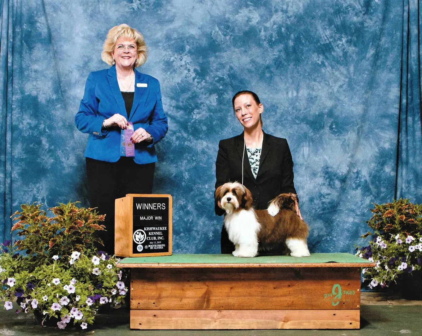Two women standing next to a small dog at a Havanese dog show.