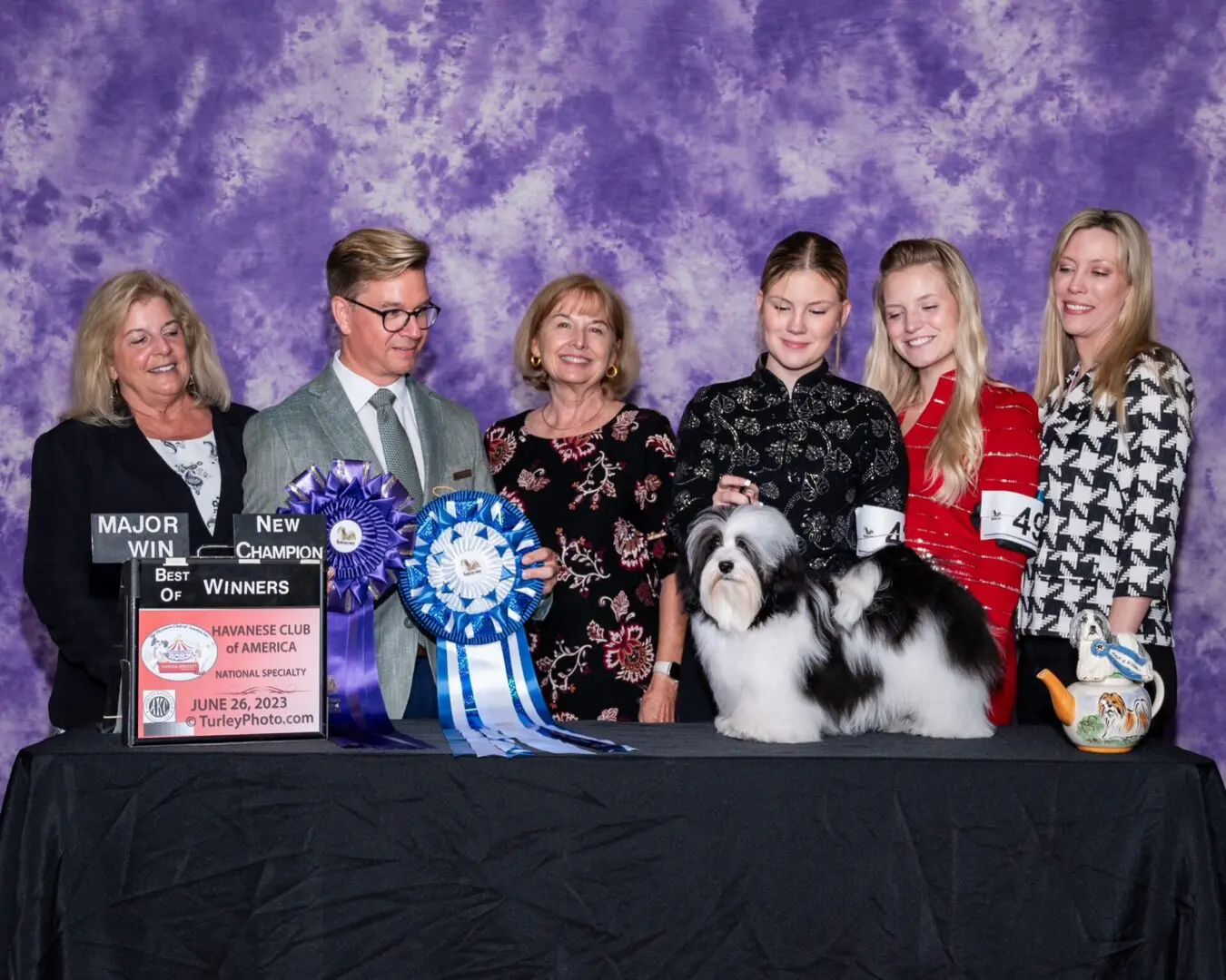 A group of people posing with a Havanese dog and ribbon, showcasing AKC Champions.